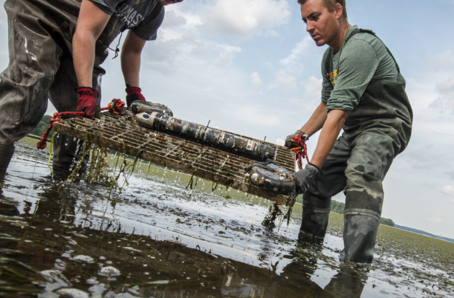Two George Mason University Flood Hazard Research Lab team members in waders retrieving water monitoring equipment from a shallow waterway during flood hazards field research