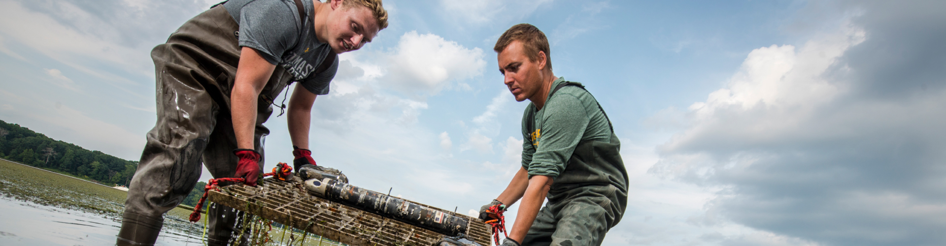 Wide-format banner of two GMU Flood Hazard Research Lab team members in waders retrieving flood monitoring equipment from a shallow waterway, used on the Flood Hazards Research Lab website