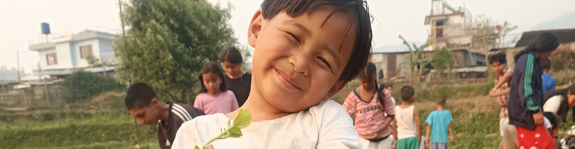 Wide-format banner of a smiling young Nepali child holding a small plant during an outdoor garden activity at Ama Ghar in Nepal