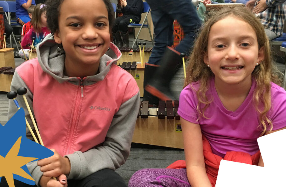 Two smiling elementary-age girls sit at Suzuki xylophones during a school music class, with other students playing instruments in the background.