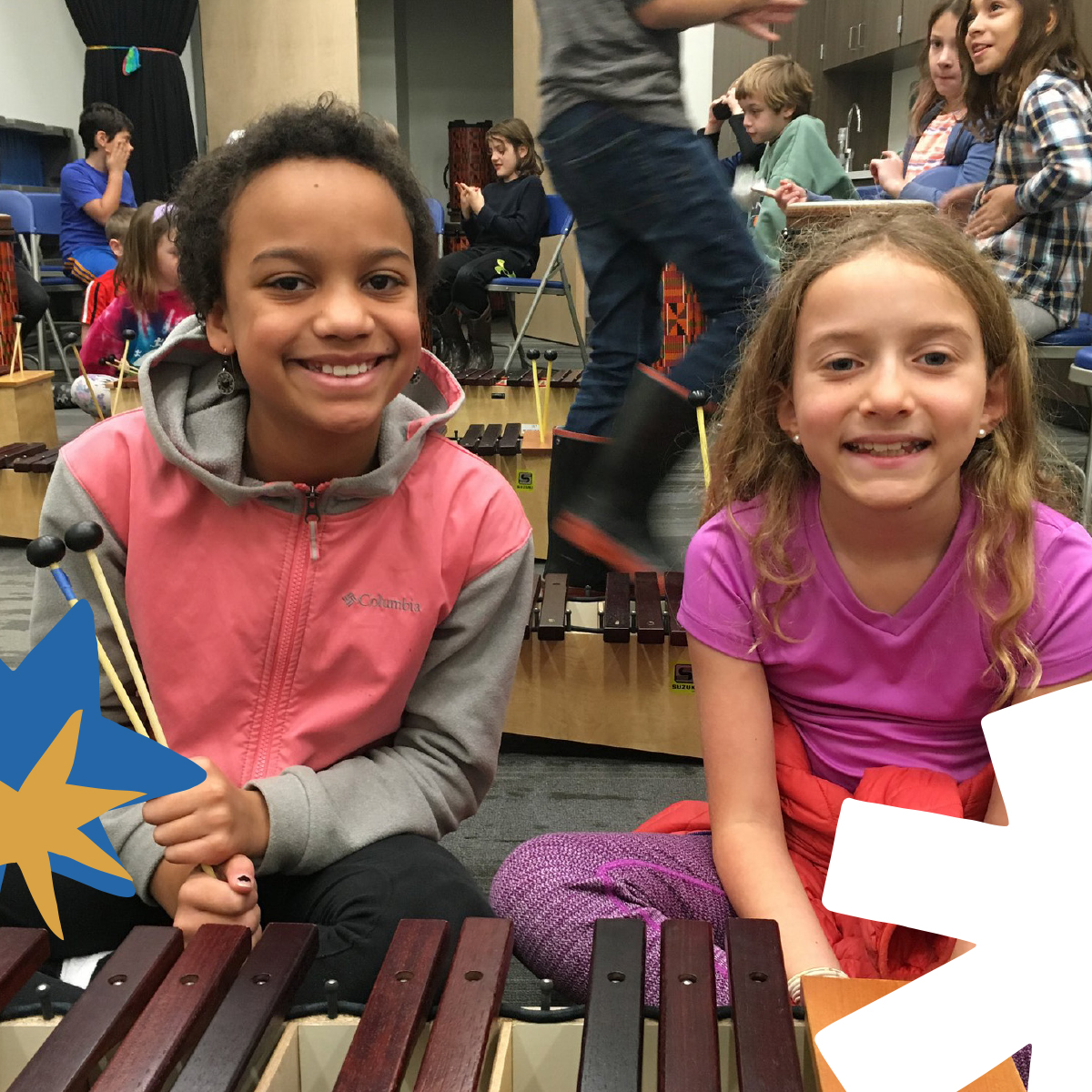 Two smiling elementary-age girls sit at Suzuki xylophones during a school music class, with other students playing instruments in the background.