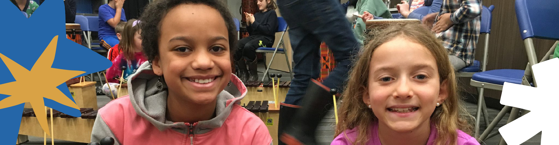 Wide banner image of two smiling elementary-age girls seated at xylophones in a music classroom, with colorful star graphics overlaid on the left and right edges.