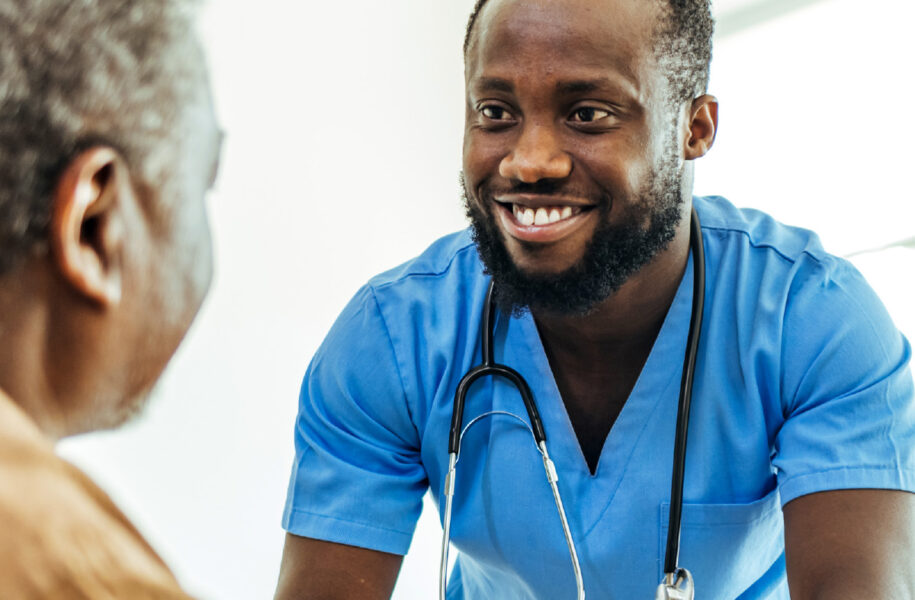 Square portfolio image of a smiling healthcare worker in blue scrubs with a stethoscope engaging warmly with a patient, representing the human-centered focus of the RUHS workforce development report